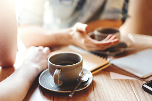 2 people at a coffee shop, drinking black coffee and one using hand gestures.