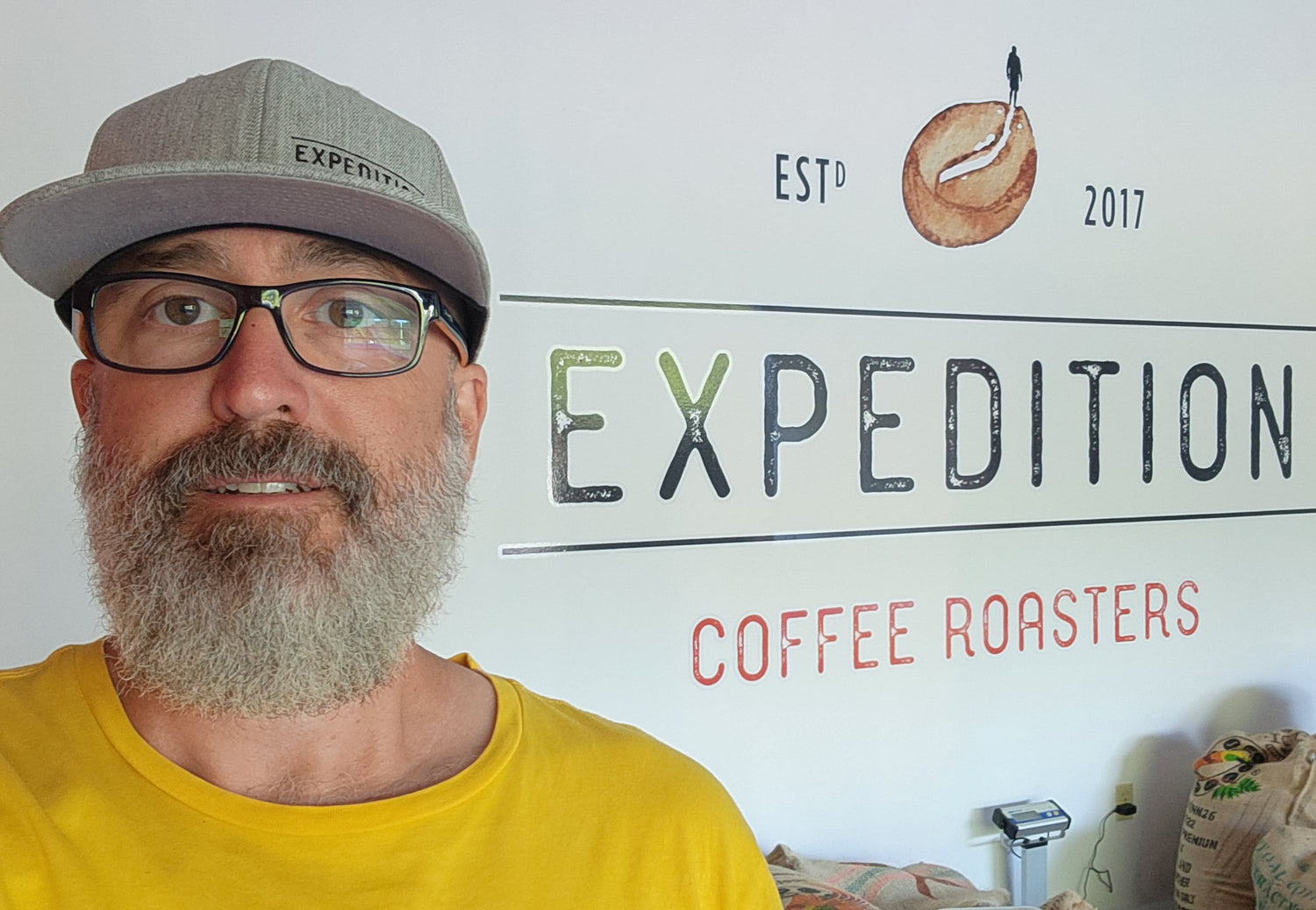 Aaron Grimeau owner and roaster of Expedition Coffee Roasters in front of his logo wall in Falmouth