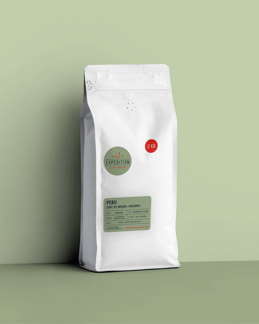 A white stand-up coffee bag with a resealable zipper, standing on a matching light green surface against a pale green background. The bag has a green circular Expedition logo, a red circular '2 KG' weight badge, and a green label reading 'PERU CAFE DE MUJER - ORGANIC'.